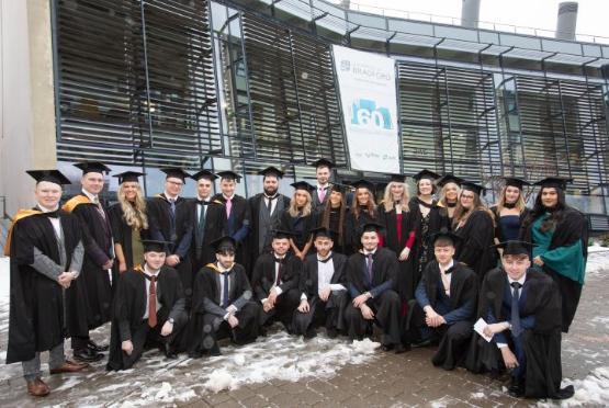 A large group of students dressed in graduation gowns pose for photo outside university building