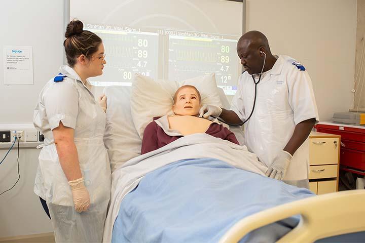 Two nursing students measuring the vitals of a medical mannequin on a hospital bed.