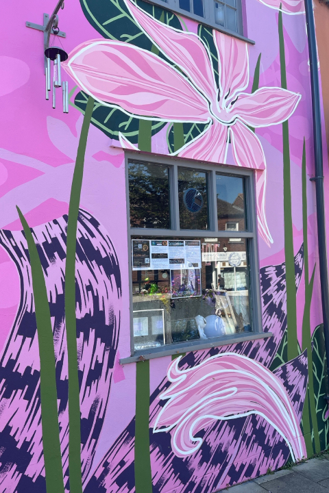 The side of a building painted pink with floral designs and a window displaying flyers.