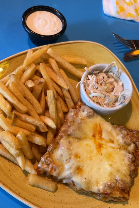 A yellow plate holding lasagne, chips and a bowl of coleslaw sits on a blue surface.