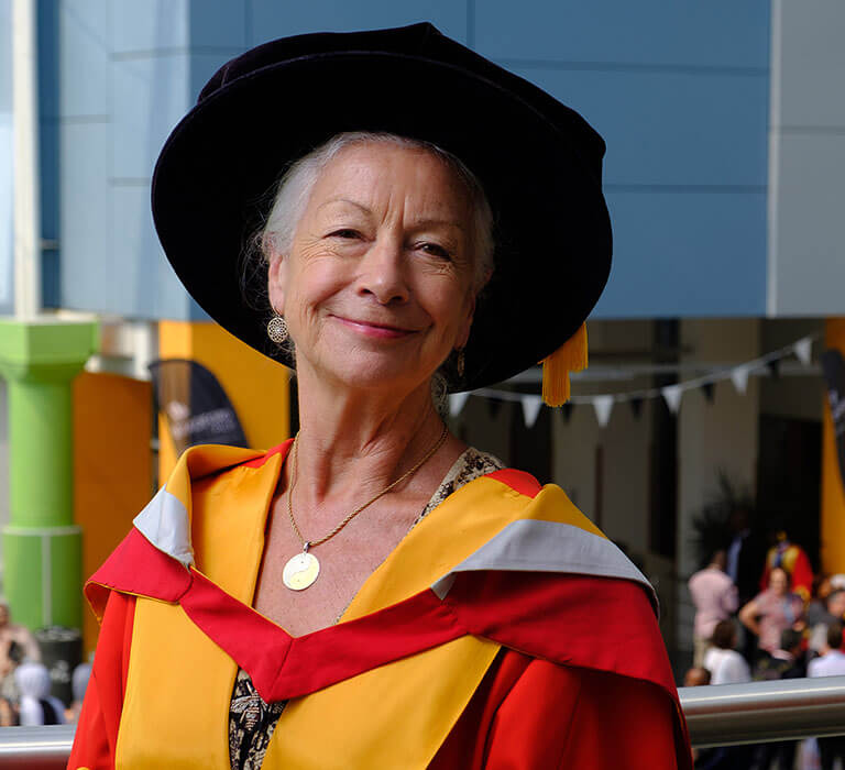 Dr Scillia Elworthy wearing her honorary graduate gown and hat while posing in the atrium of the Richmond Building.