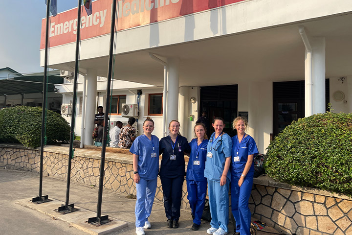 A group of student nurses wearing their scrubs smile at the camera outside of a hospital in the blazing sunshine