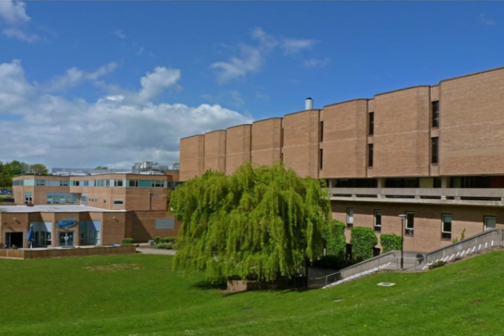 A large, brown university appears behind a grassy field with several trees.