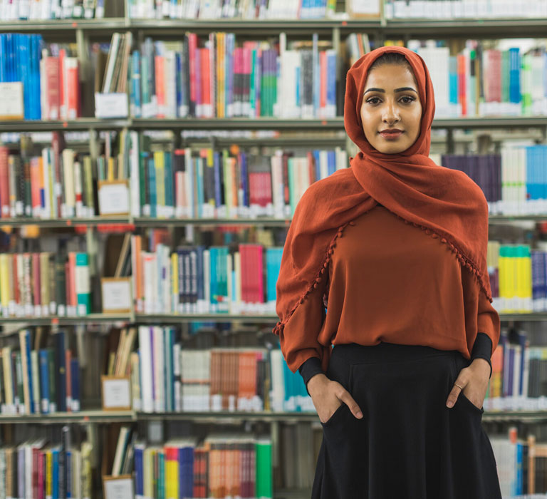 A student smiling at the camera standing in front of library shelves filled with books