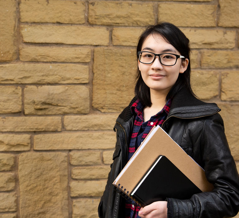 A student holding notebooks standing next to a brick wall looking at the camera