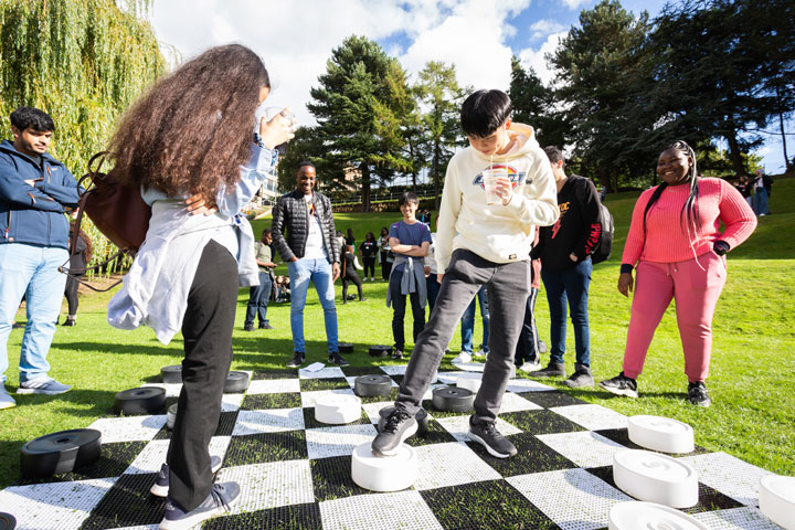 Two students playing draughts on an oversized board on a lawn with other students watching