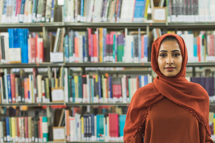 A student smiling at the camera standing in front of library shelves filled with books