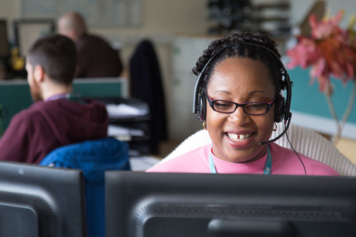 A person is sitting in front of a computer monitor wearing a phone headset and smiling