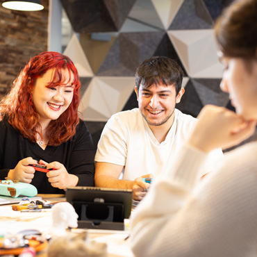 Two students playing on a switch console at a table in a shared student space as another student is using art supplies in the foreground.