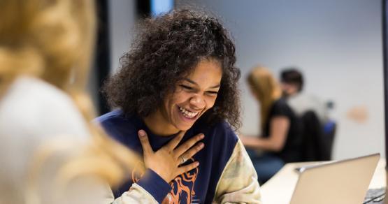 A student smiling while studying on a laptop in a shared study space on campus.