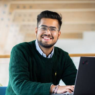 A man wearing smart clothes and glasses smiles while sat at a desk.