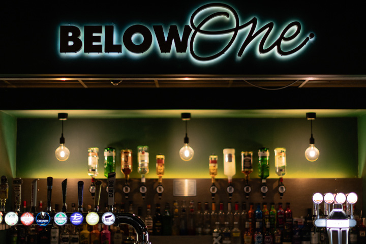 The bar at Below One, a neon sign displays the words Below One and a selection of alcoholic beverages can be seen on tap and behind the bar.