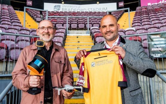 One person holds a small drone camera while a person stood next to them holds a football shirt as they both stand together in a football stadium