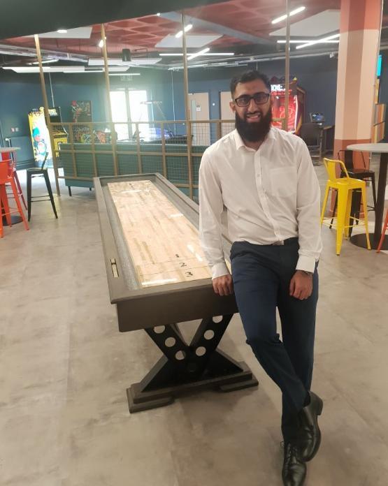 A university staff member stands to the side of a wooden shuffleboard table inside a students' union room