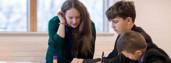 A teacher stands over between two school pupils who are sat at a wooden desk