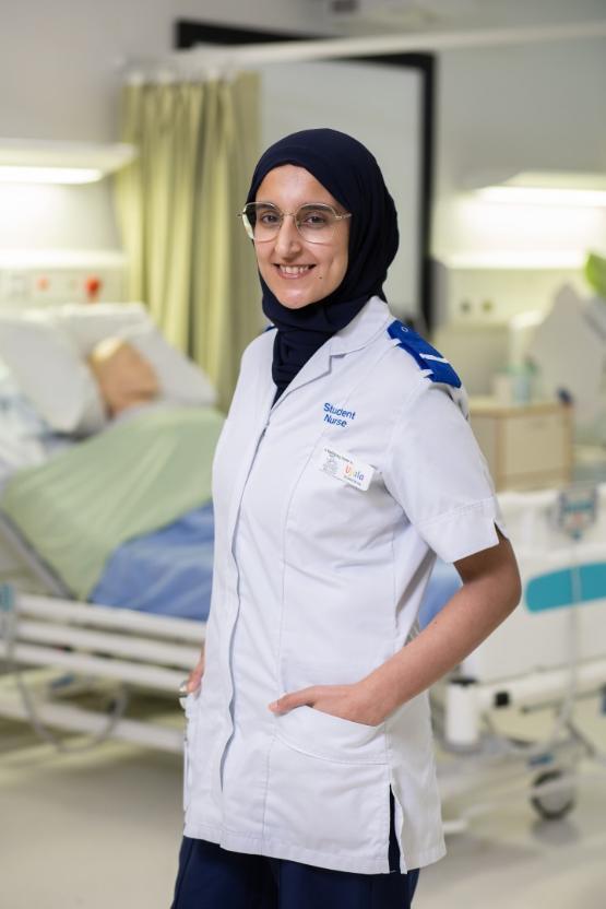 A university student stands up in front of a bed in a mock hospital ward wearing a uniform with student nurse written on it