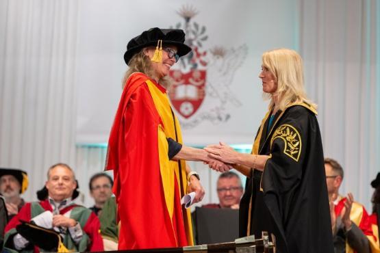 a person in graduation gown shake hands with another person on stage during graduation ceremony