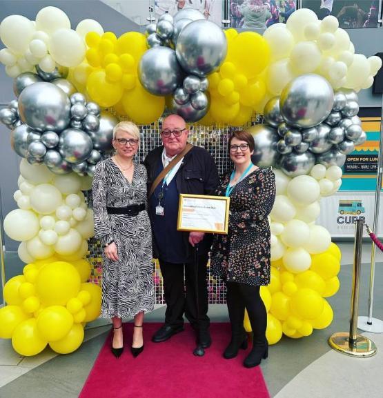 Three people stood up on a red carpet in front of a large balloon arch at an awards ceremony