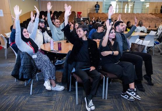 A group of students sat down at a table each with their hands in the air and smiling at a conference