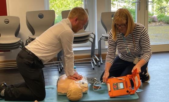 One person using CPR techniques on a dummy as a person next to them holds a defibrillator