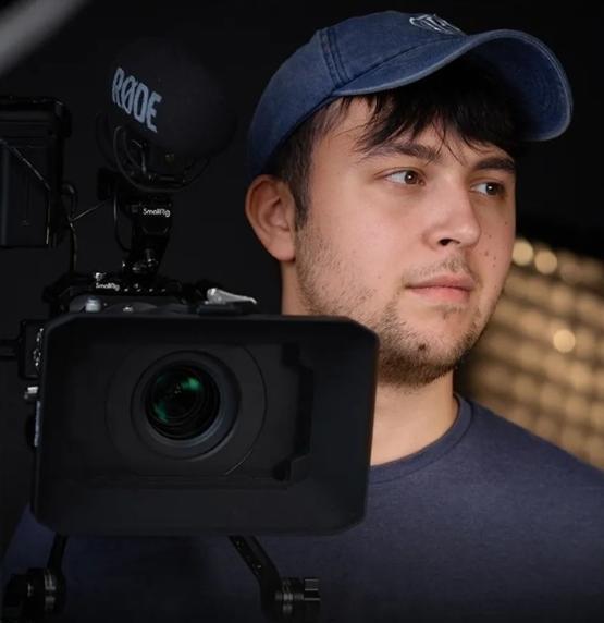 former student wearing baseball cap holding a TV camera over their right shoulder