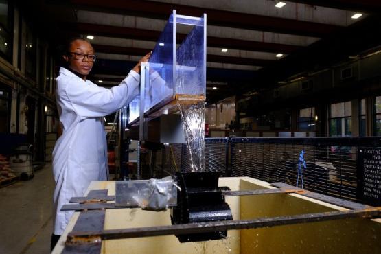 A university student in a white coat and protective glasses stands up and carries out an experiment with water dripping off the end of a plastic box