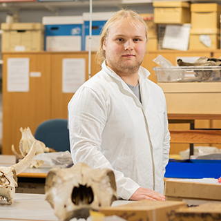 A student in the archaeology laboratory at the university standing proudly in a white coat smiling at the camera