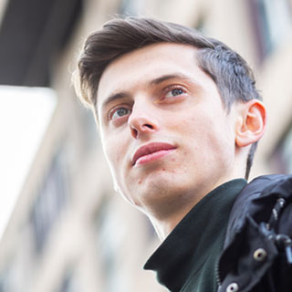 A student wearing a black coat standing outside in front of a large brick building.