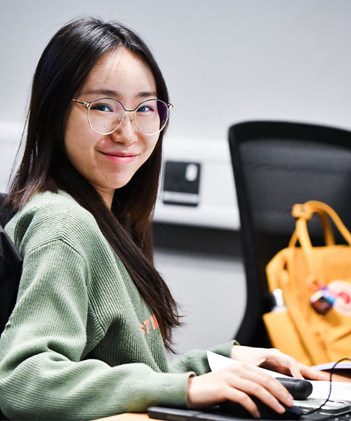 A student working on a computer, smiling at the camera.